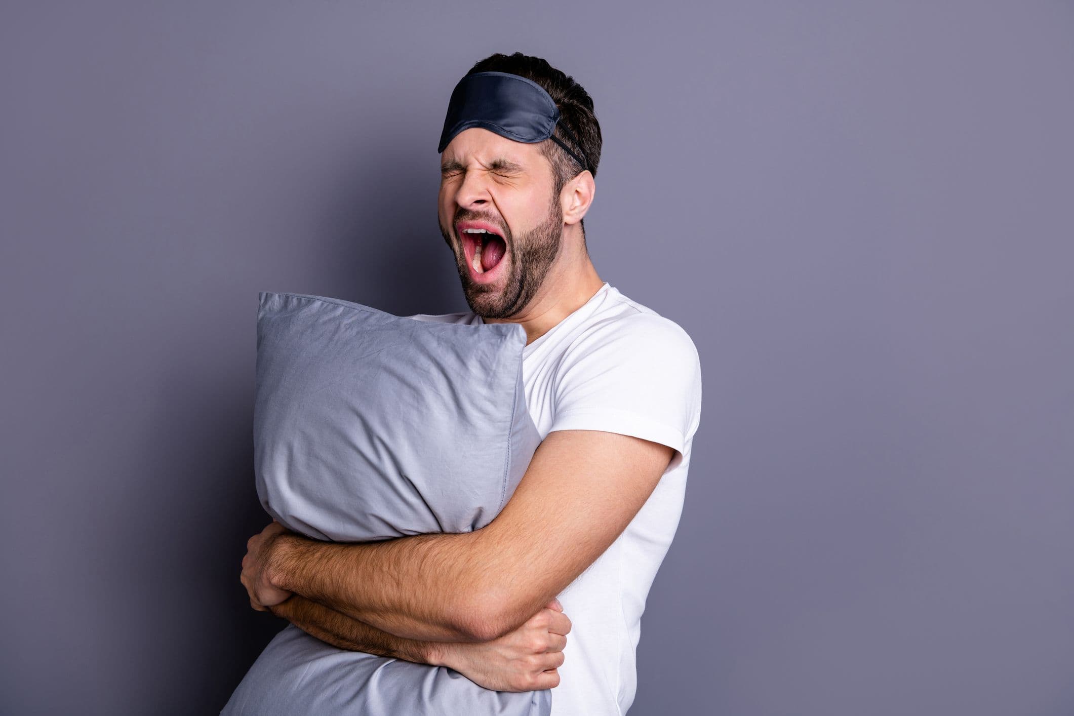 A man looking tired and yawning while holding a pillow and wearing an eyemask
