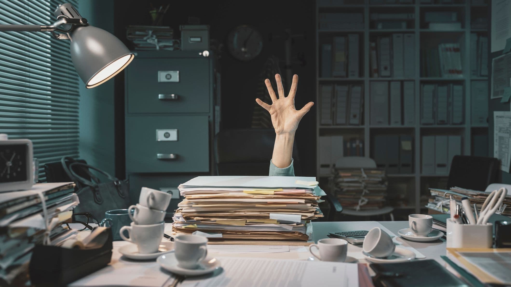 Stressad person som ligger på golvet och sträcker sig bakom en hög med arbetsuppgifter och kaffekoppar