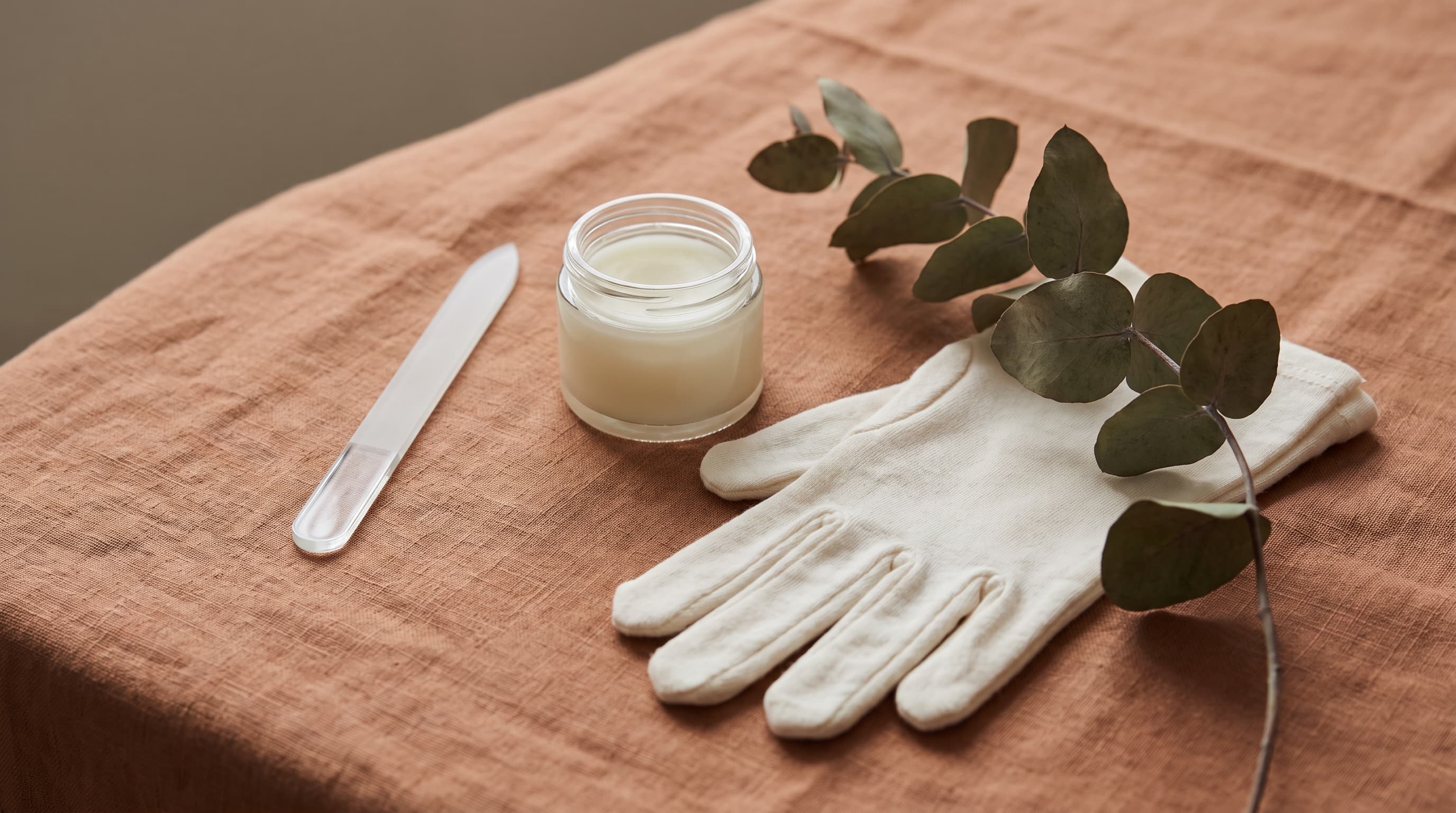 A glass nail file, a small jar of hand cream and a pair of white cotton gloves with a eucalyptus sprig on a terracotta linen cloth